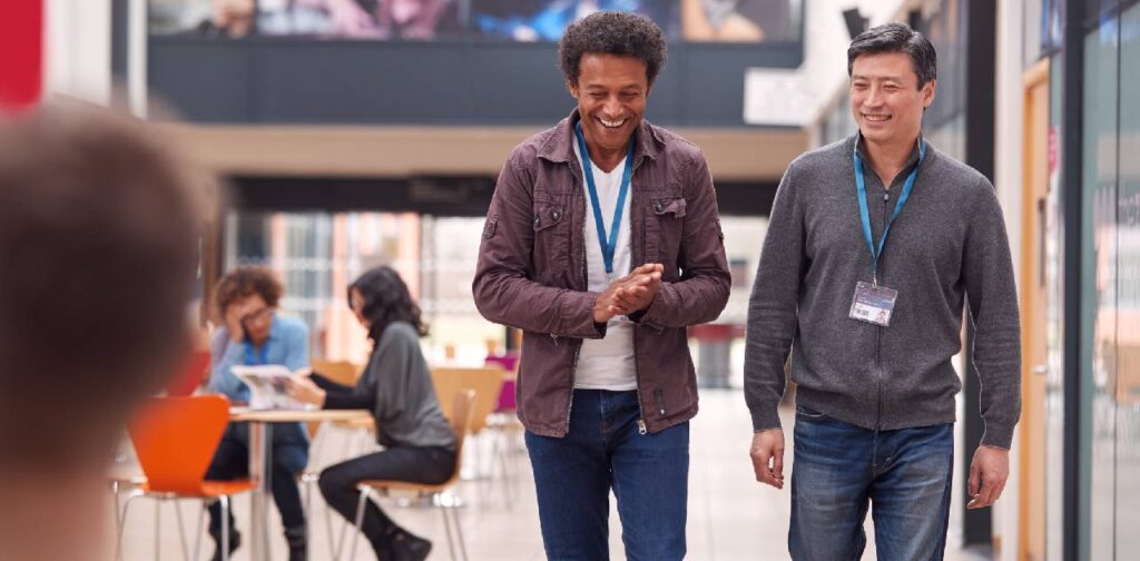 Two Mature Male Students Or Teachers Walking Through Communal Hall Of Busy College Campus Building